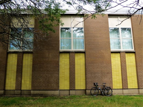 A tour bike leaning on a mid century modern school wall with three yellow brick vertical highlights below three vertical windows.