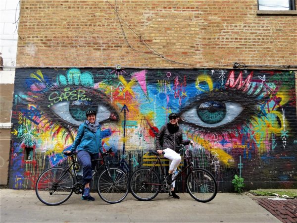 Two CBA bike tour riders leaning on a multi-colored mural of two feminine eyes