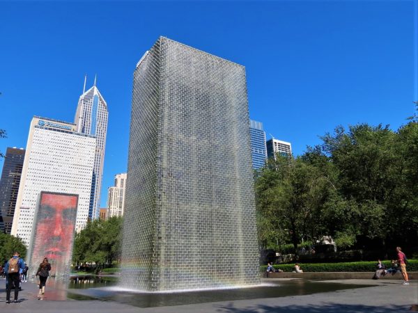 A CBA bike tour rider taking a picture of two vertical glass block structures releasing water, one can been seen to have a human face, with skyscrapers in the background.