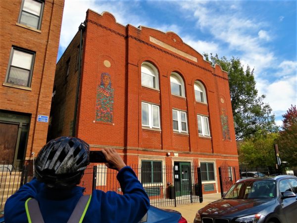 A bike tour rider taking a picture of a three story red brick former synagogue with painted portraits at the corners.