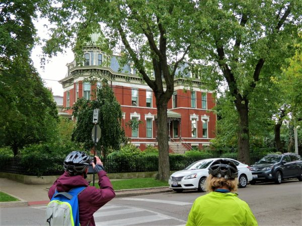 Two CBA bike tour riders looking at a four story red brick grey roofed Second Empire mansion.