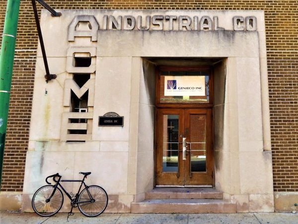 A tour bike leaning on the Art Deco stone business facade that reads ACME vertically to the left of the door and INDUSTRIAL above it.