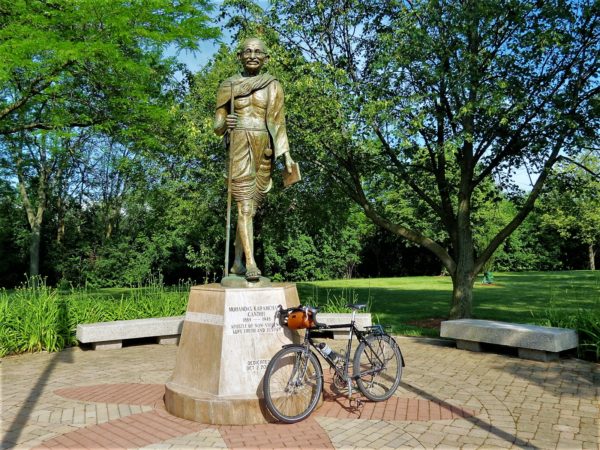 A tour bike leaning on the base of a bronze statue of Gandhi in a short dhoti and glasses holding a walking stick.
