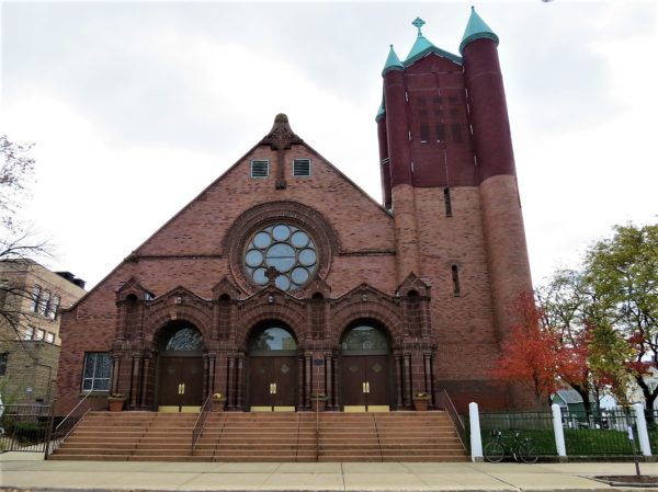 A tour bike standing in front of a low steep roofed red brick church with two toned red brick patina copper topped tour.