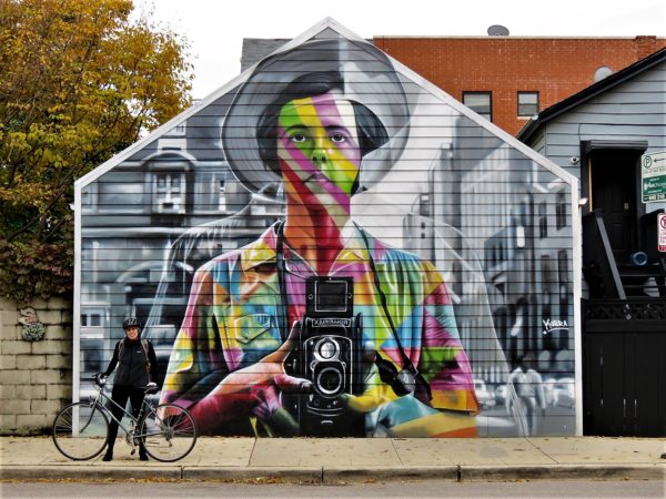 A CBA bike tour rider standing nest to a garage mural of a rainbow cl,olored woman in a wide brim hat holding a vintage top view finder camera on a black and white background