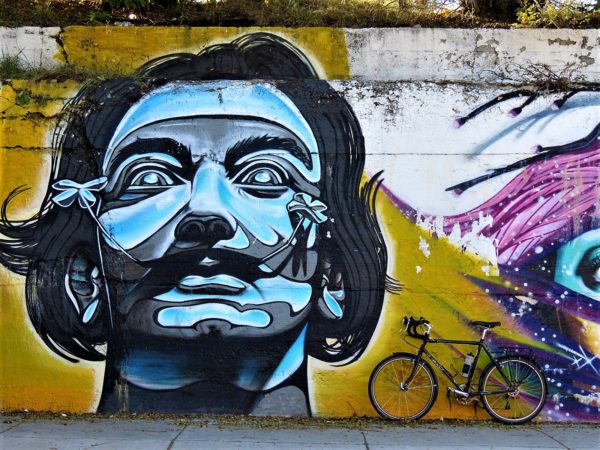 A tour bike leaning on a yellow painted wall with a aqua and black street portrait of Salvadore Dali with wide mustache and his head cocked back.