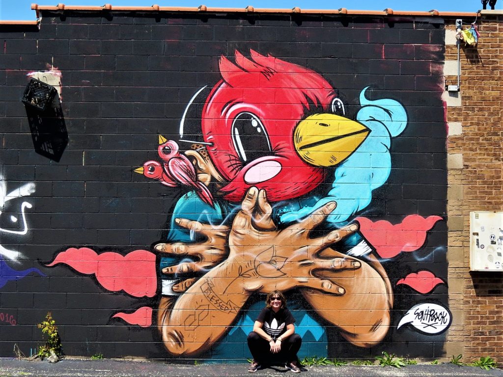 A CBA bike tour rider squatting below a mural of a figure with hands crossed wearing a red bird mask,