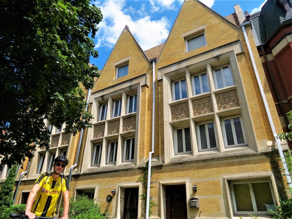 A CBA bike tour rider in font of a three story yellow brick row houses with two bands of windows.