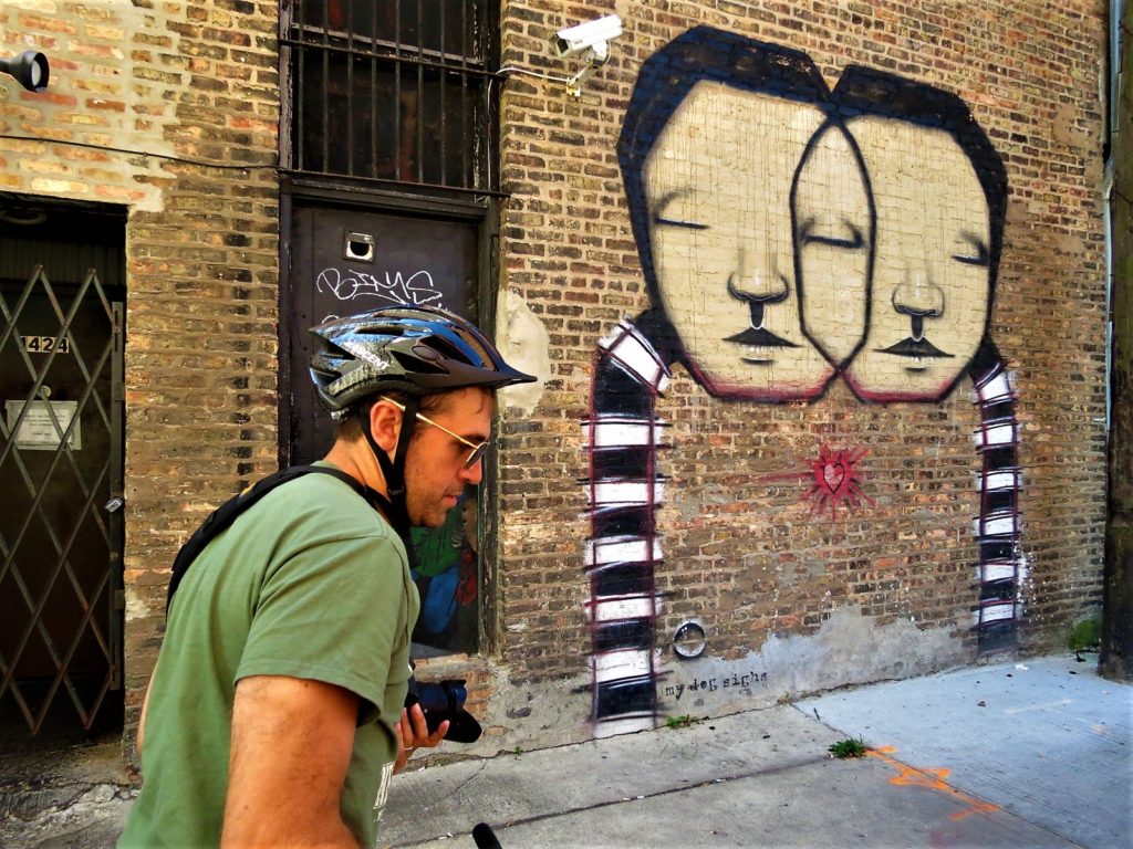 A CBA bike tour rider in front of a mural of two human faces sgaring one eye and a black and white scarf like object