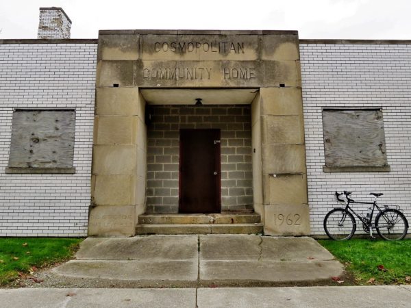 A tpur bike leaning to the side of a stone entry way with the above door epigram Cosmopolitan Community Home with boarded up windows on either side