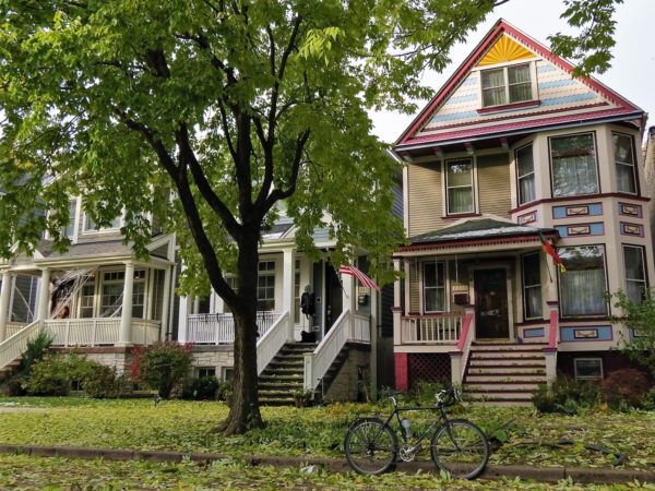 A tour bike standing in front of a three story wood fram home with a bay window that extends from the first and second floors, painted in light blue, purple, and pink colors and sun rays in the point of the gable.