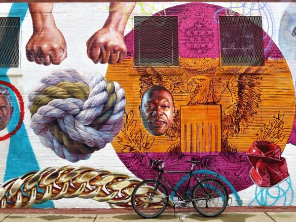 A tour bicycle leaning on a mural of a knot, two fists, a man;s face and a pink and orange circle with the American eagle inside