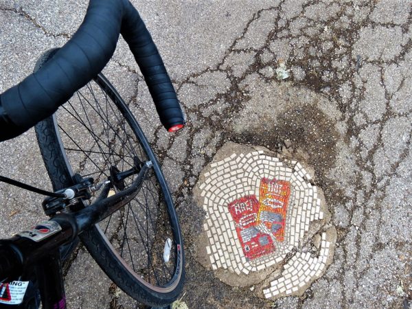 A tour bicycle front wheel next to a mosaic constructed in the street of two chip bags.