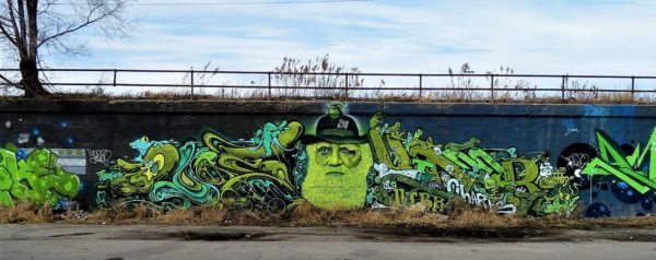A tour bike leaning on a green mural with heavy detail on either side of a bearded man in a hat on a black wall