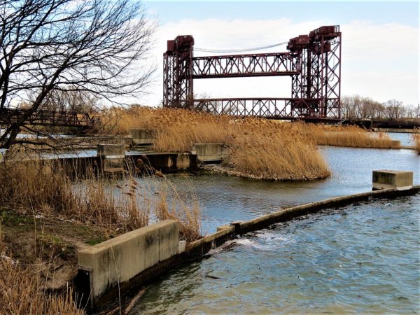 A stepped water structure surrounded by brown tall grass with two vertical lift bridges in the background.