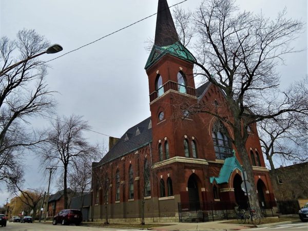 A tour bike standing in at the corner of a high pitched red brick one tower former church.