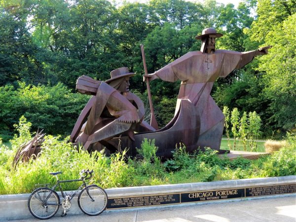 A tour bike standing next to three brown metal figures in a boat with a priest pointing into the distance and a Native person looking down pulling the boat.