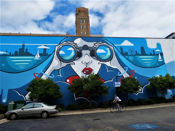 A CBA bike tour rider cycling passed a blue based mural of the Chicago skyline on either side of a girl looking forward through binoculars