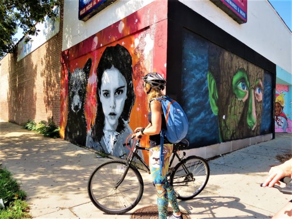 A CBA bike tpour rider standing at a corner looking at two murals, one an orange mural with black and white child and baby bear, the other a male face covered in bark except for eyes and nose