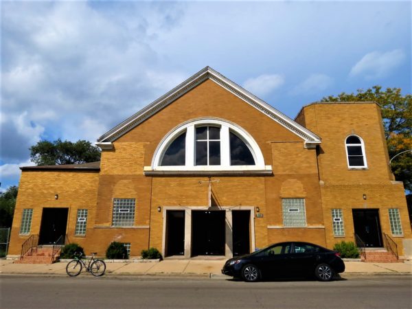 A tour bike standing n ront of a two story yellow brick high pitched roof former synagogue with a semi circle middle window and a short side tower.