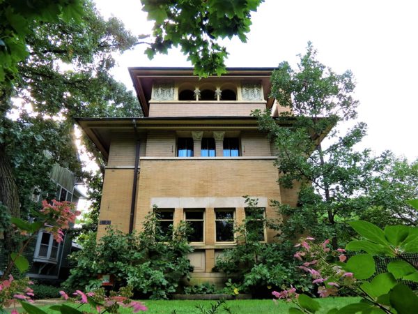 A three story Prairie Style home shot through the green leaves of trees and bushes