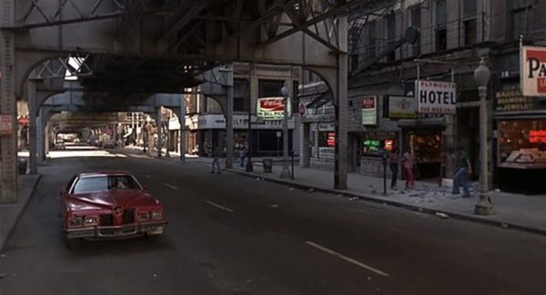 A Blues Brothers movie still of a car driving underneath elevated rail track passing storefronts and a hotel