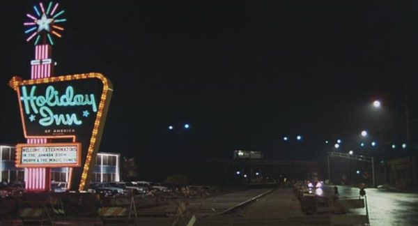 A Blues Brothers movie still of the Bluesmobile approacing a large neon Holiday Inn sign