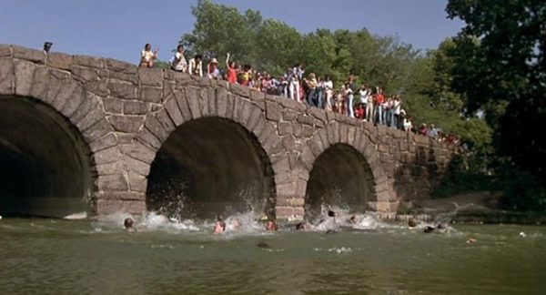 A Blues Brothers movie still of people on a stone three arch bridge looking down at others thrashing in the water.