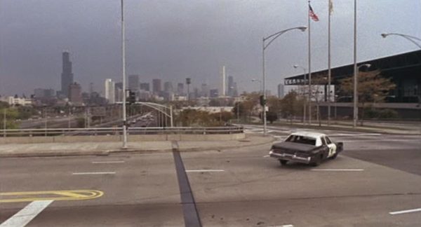 A Blues Brothers movie still of the Bluesmobile making a left turn with the 1980s Chicago skyline in the distance.