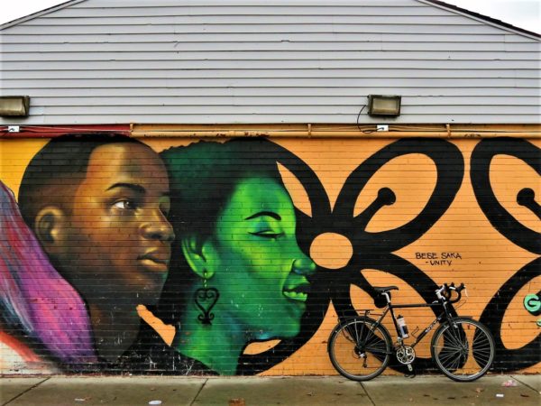 A tour bike leaning on a mural with an African American man and woman, stylistically painted in green, facing right on a yellow background patterned with a black outline flower