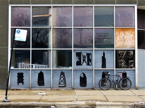 A tour bike leaning on a four by six square pane window with the bottom six panels filled with silhouettes of buildings and objects