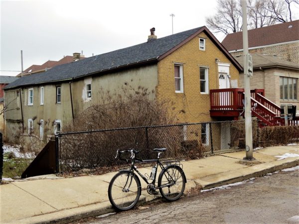 A tour bike standing in front of a long two story home, with the first floor below street grade and a wood stairs up to the second floor entrance