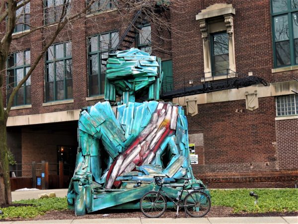 A tour bicycle standing in front of a Buddha figure made of wood painted aqua blue with a red sash siting, a brick building behind