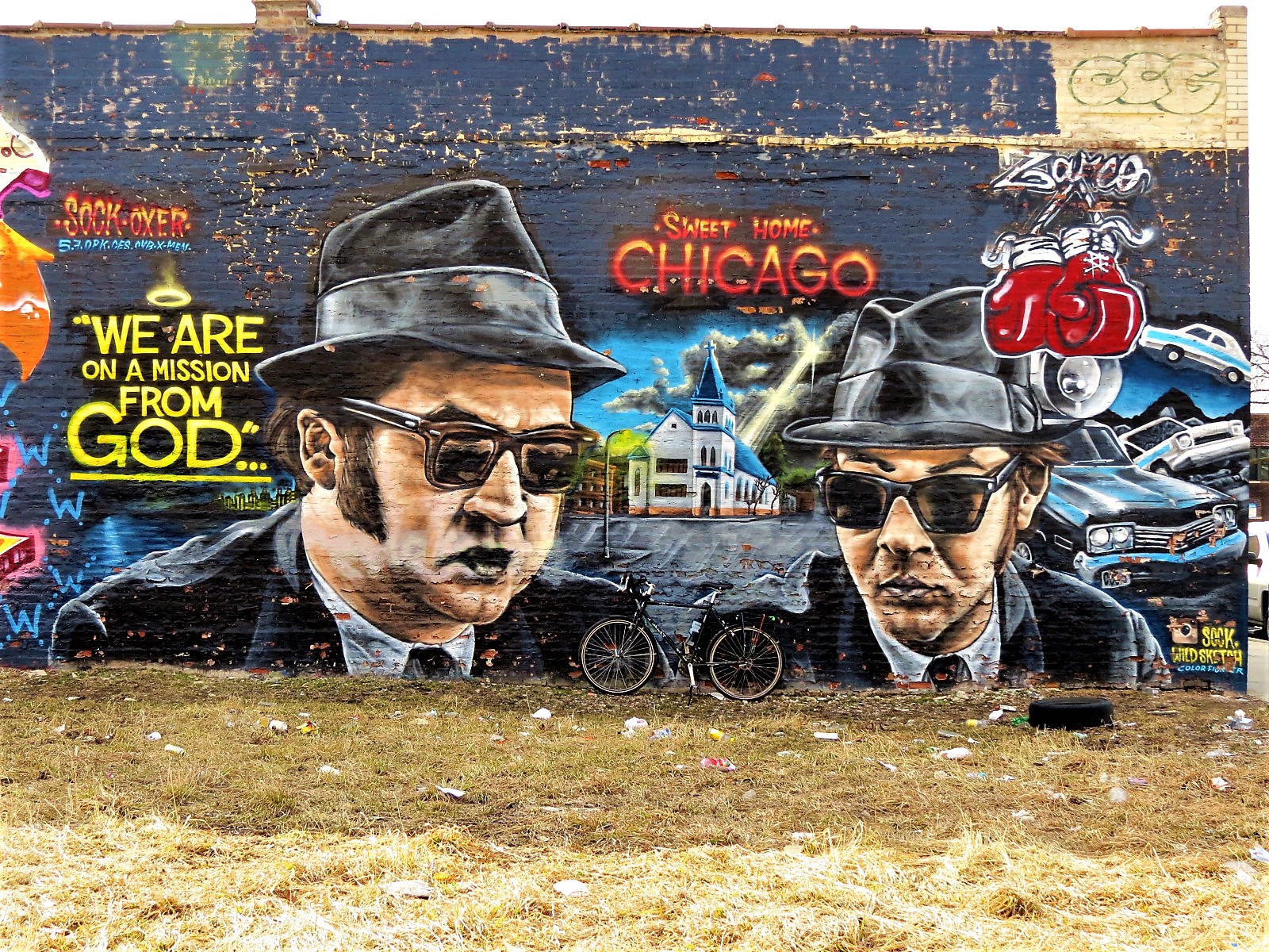 A tour bike leaning on a mural of the Blues Brothers and yellow text that reads We are on a mission from God.