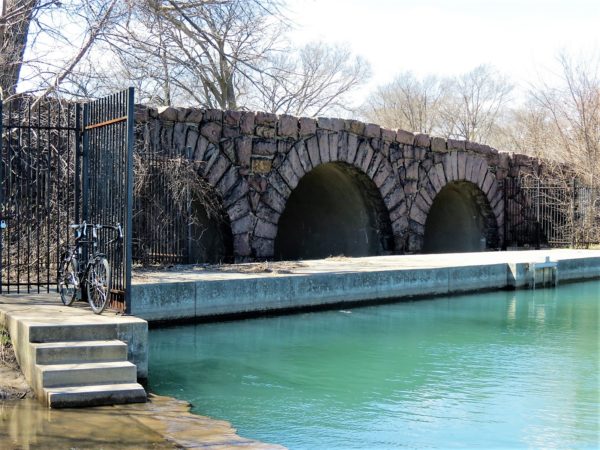 A tur bike standing next to a hree arch stone bridge over cool blue green water.