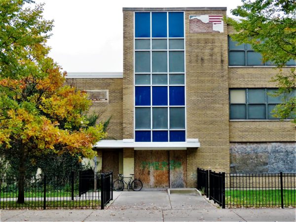 A tour bike leaning next to the boarded entrance to a closed school with a large blue and gray vertical window.