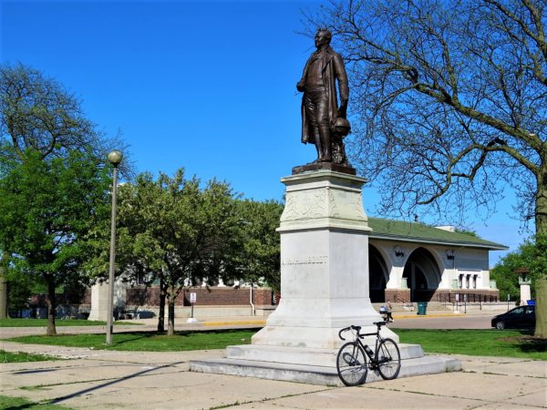 A tour bike leaning on the white stone pedestal of a bronze male statue with a Prairie School style building in the background.