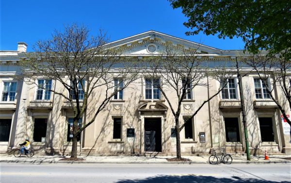 A tour bike standing in front of a Classical Revival white stone former bank building with a cyclist riding passed.