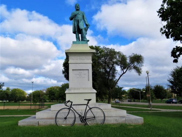 A tour bike leaning on the white stone pedestal of a green bronze standing man staute,