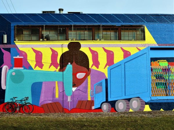 A tour bike leaning on a flat cartoon style bright mural of a woman of color working behind a sewing machine