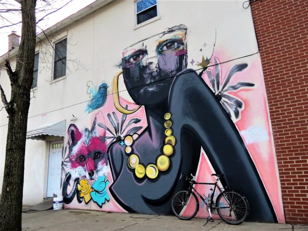 A tour bike leaningon a collaboration mural of a woman with a flat cartoon style body and big pearl necklace with realistic face.