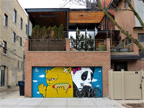 A tour bike leaning on a garage door with a murtal of a light brown bear with hotdog in hand with back to a panda colored bear with a red bow.