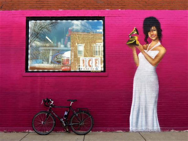 a tour bike leaning on a magenta painted wall with the figure of the deceased Tejano singer Selena in a white dress holding up a Grammy,