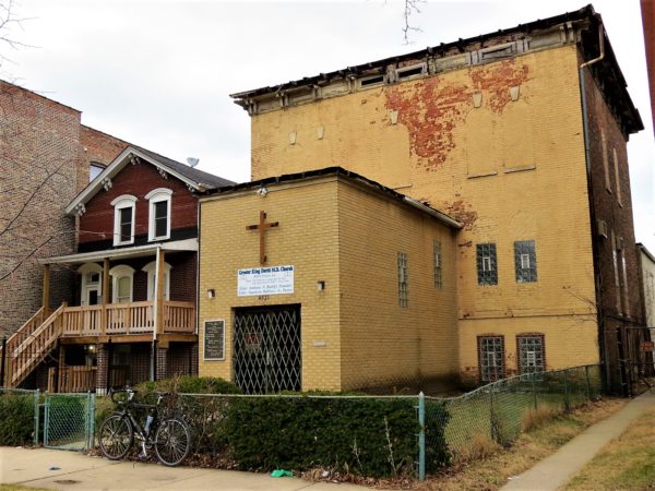 A tour bike leaning on the fence in front of a flat roof Italiante three story converted to church with painted yellow brick and a font entry extension, next door to a two and half story 1870 sloped roof Italianate residence with wooden stairs up to a second floor entry