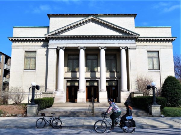 A tour bike standing in fonr of a Classical Revival church building as a man and woman on one bike ride by.