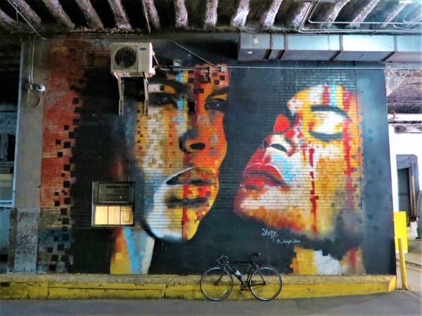 In a parking garage a tour bike is leaning in front of a mural of an African American man and woman's portraits done in splashes of light blues yellow, and red
