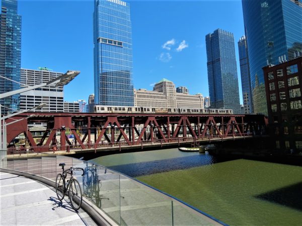 A tour bike leaning in the foreground above a greenish river spanned by a double deck maroon painted movable bridge with an elevated train on its top deck.