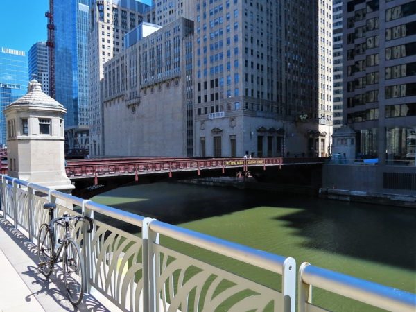 A tour bike leaning in the foreground with a painted maroon deck truss movable bridge spanning greenish water with two stone tender houses on either end and a smooth faced Art Deco Opera building in the background