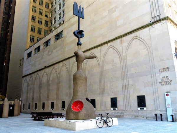 A tour bike standing at the base of an abstract sculpture with two appendage like extensions, a red pockmark and a trident coming from the top.