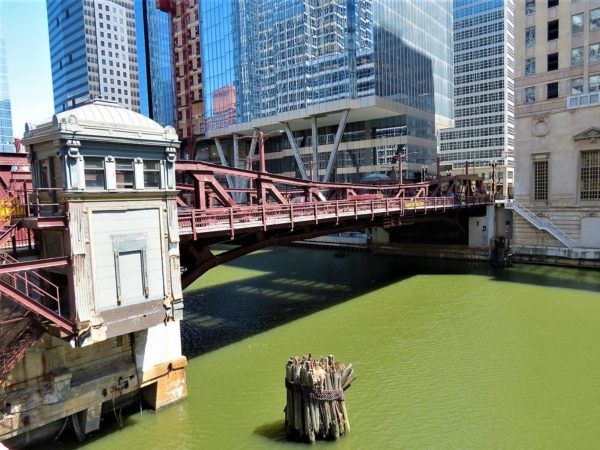 A copper and white minder house to the left of a painted maroon pony truss movable bridge with the base of a skyscraper behind.